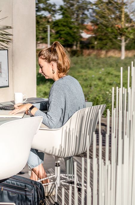 Eine Frau sitzt draußen an einem Schreibtisch und arbeitet an einem Computer. Sie trägt einen grauen Pullover und hat ihr Haar zu einem Pferdeschwanz gebunden. In der Nähe steht ein weißer Becher, umgeben von Grün - ein inspirierender Ort für die Arbeit im Garten- und Landschaftsbau.