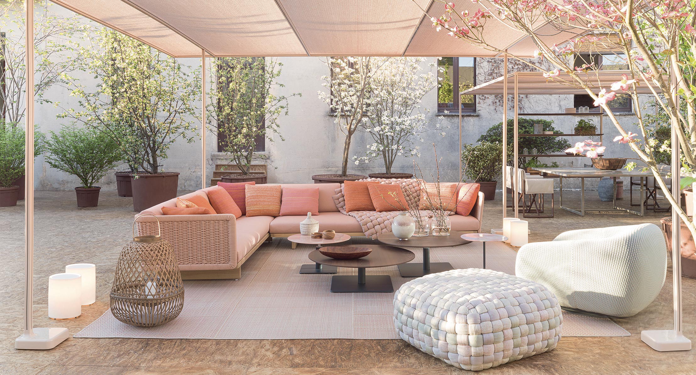 Moderne Außenterrasse mit rosafarbenem Sektionssofa, Poufs, Couchtischen, Laternen und blühenden Bäumen im Hintergrund - ein Beispiel für die Inspiration von Burelbach Gardendesign aus Deutschland.
