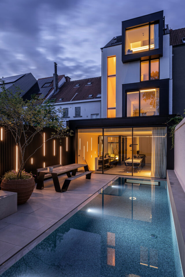 Moderner städtischer Hinterhof in der Abenddämmerung mit einem beleuchteten rechteckigen Swimmingpool, stilvollen Gartenmöbeln, Topfpflanzen und einem modernen mehrstöckigen Haus mit großen Fenstern und warmer Innenbeleuchtung - perfekt für den Garten- und Landschaftsbau in Deutschland und Luxemburg.