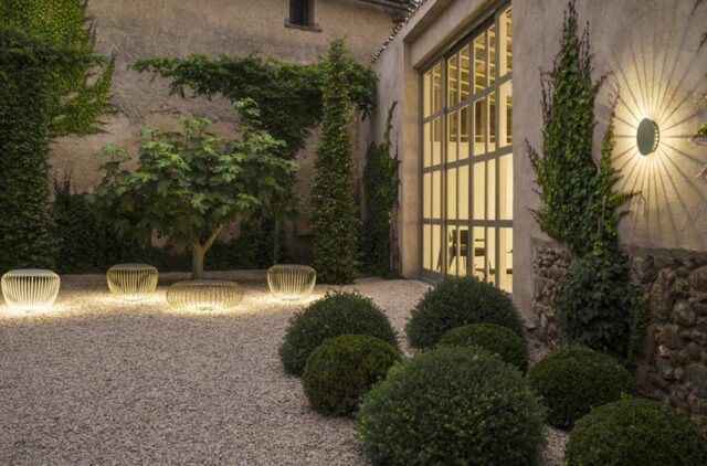 Ein friedlicher Innenhof mit Kiesboden, Rundsträuchern, einem kleinen Baum und mit Weinreben bewachsenen Mauern. Moderne kreisförmige Außenleuchten leuchten sanft und betonen das handwerklich inspirierte Gartendesign und die eleganten Gartenmöbel durch Glastüren mit einem einladenden Innenraum, der von innen sichtbar ist.