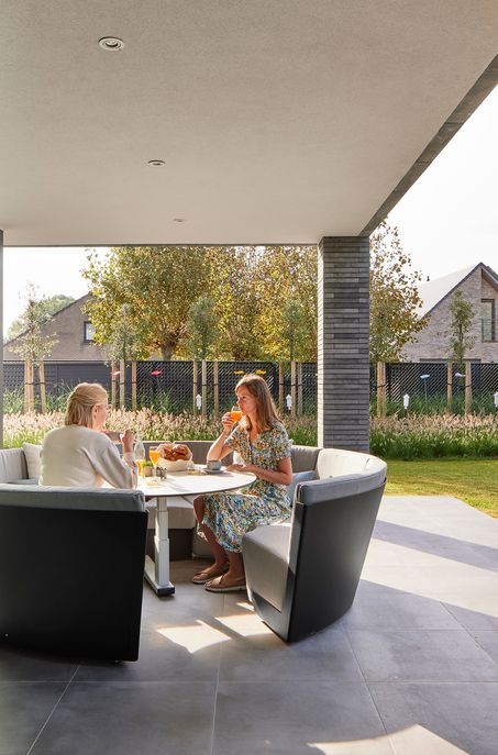 Zwei Frauen sitzen an einem runden Tisch im Freien auf einer überdachten Terrasse und genießen Getränke und Snacks. Der Gartenentwurf von Burelbach zeigt grünes Gras und moderne Häuser im Hintergrund unter einem sonnigen Himmel.