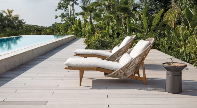 Zwei gepolsterte Loungesessel von Burelbach Handwerk stehen auf einem Holzdeck neben einem Pool, umgeben von üppigen tropischen Pflanzen. Ein kleiner Beistelltisch mit einem Glas und einer Karaffe sorgt für Komfort - ganz im Stil von Deutschland und Luxemburg.