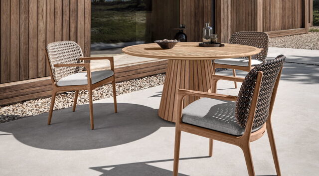 Ein modernes Gartenmöbel-Terrassenset mit einem runden Holztisch und drei gepolsterten Stühlen mit geflochtener Rückenlehne, die auf einer Betonfläche neben einem holzverkleideten Gebäude stehen - perfekt für Häuser in Deutschland und Luxemburg.