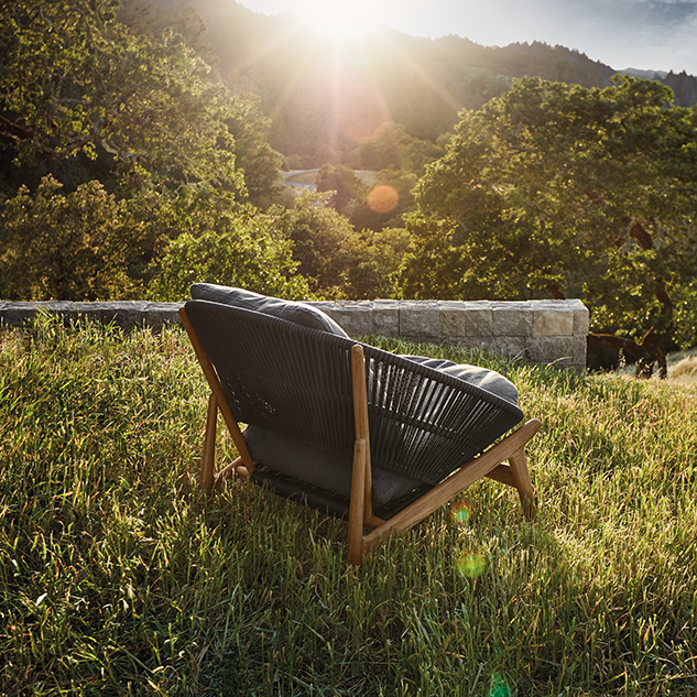 Ein moderner Outdoor-Stuhl mit dunklen Polstern steht auf einem Grashügel vor einer Steinmauer und Bäumen und zeigt exquisites Gardendesign, während das Sonnenlicht durch den Hintergrund fällt.