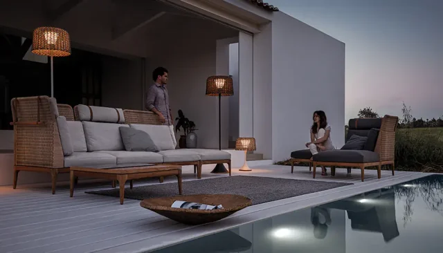 Ein Mann und eine Frau entspannen sich auf Burelbach Gartenmöbeln neben einem modernen Haus in der Abenddämmerung, mit sanfter Beleuchtung und einem Pool im Vordergrund.