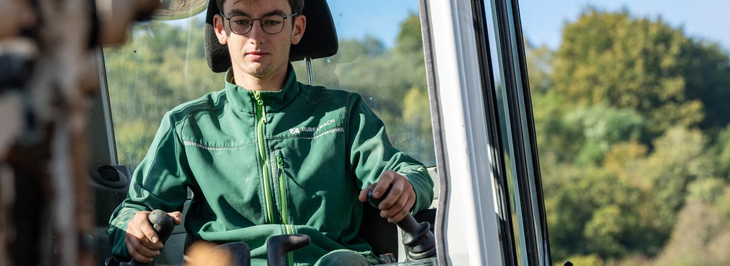 Eine Person mit Brille und grüner Jacke bedient Maschinen im Freien, die das Handwerk im Garten- und Landschaftsbau präsentieren, während im Hintergrund Bäume und blauer Himmel zu sehen sind. Burelbachs Fachwissen ist in dieser Szene offensichtlich.