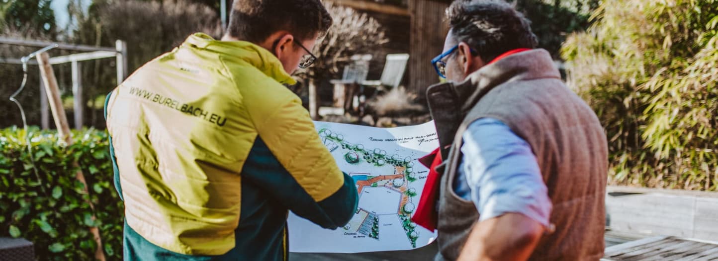 Zwei Personen stehen im Freien und diskutieren über einen Garten- und Landschaftsbau-Plan von Burelbach. Einer trägt eine gelbe Jacke mit Aufschrift, der andere eine graue Weste. Im Hintergrund sind Grünflächen, Gartenmöbel und eine Holzkonstruktion zu sehen.
