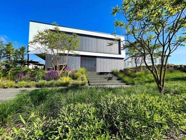 Ein modernes zweistöckiges Haus mit großen Fenstern und grauen Fensterläden, umgeben von üppig grünem Gras, Büschen und Bäumen unter einem strahlend blauen Himmel, verfügt über elegante Gartenmöbel, die das Gardendesign aufwerten. Eine Treppe führt hinauf zum einladenden Eingang.