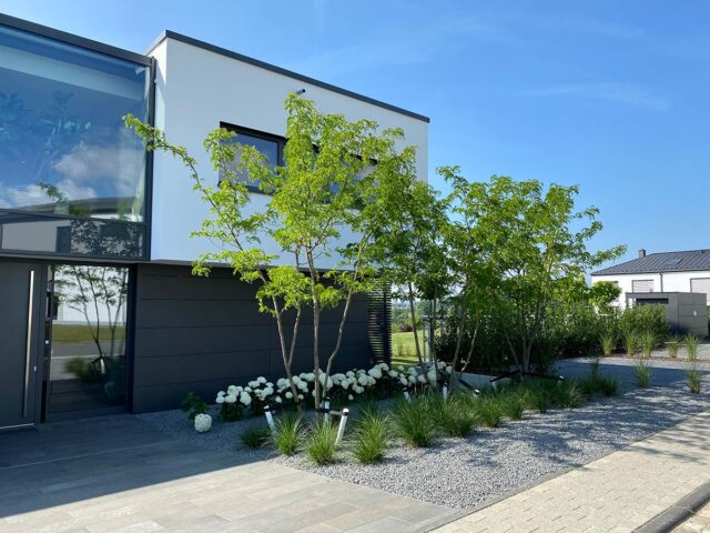 Ein modernes zweistöckiges Haus mit großen Fenstern und einer minimalistischen Gartengestaltung mit ordentlich gestutzten Pflanzen, weißen Blumen, Kiesbeeten und stilvollen Gartenmöbeln entlang eines gefliesten Weges unter dem klaren blauen Himmel.