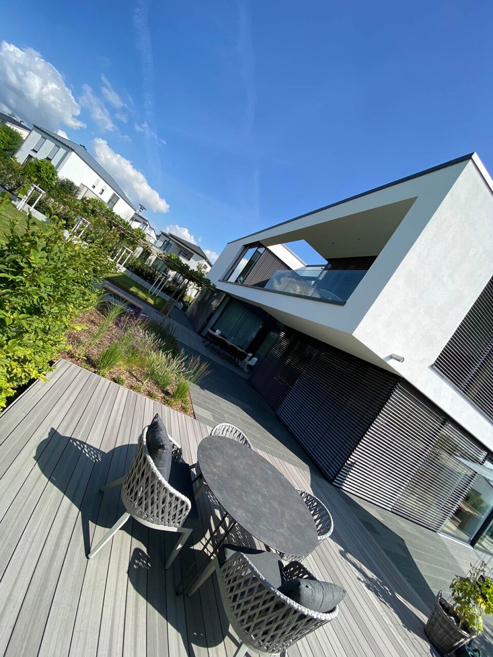 Ein modernes weißes Haus mit großen Fenstern und einem Balkon, mit stilvollen Gartenmöbeln auf einer Holzterrasse mit rundem Tisch und Stühlen, umgeben von üppigem Grün unter einem klaren blauen Himmel.