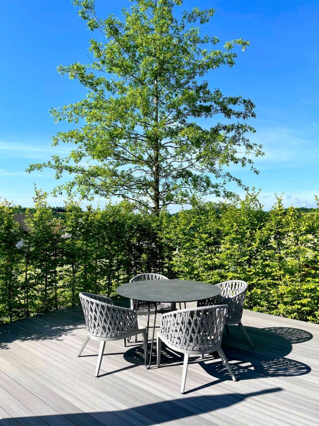 Ein runder Tisch mit vier geflochtenen Stühlen steht auf einer Holzterrasse, umgeben von üppigen grünen Büschen und einem hohen Baum, der die Handwerkskunst des Garten- und Landschaftsbaus unter einem strahlend blauen Himmel zeigt.