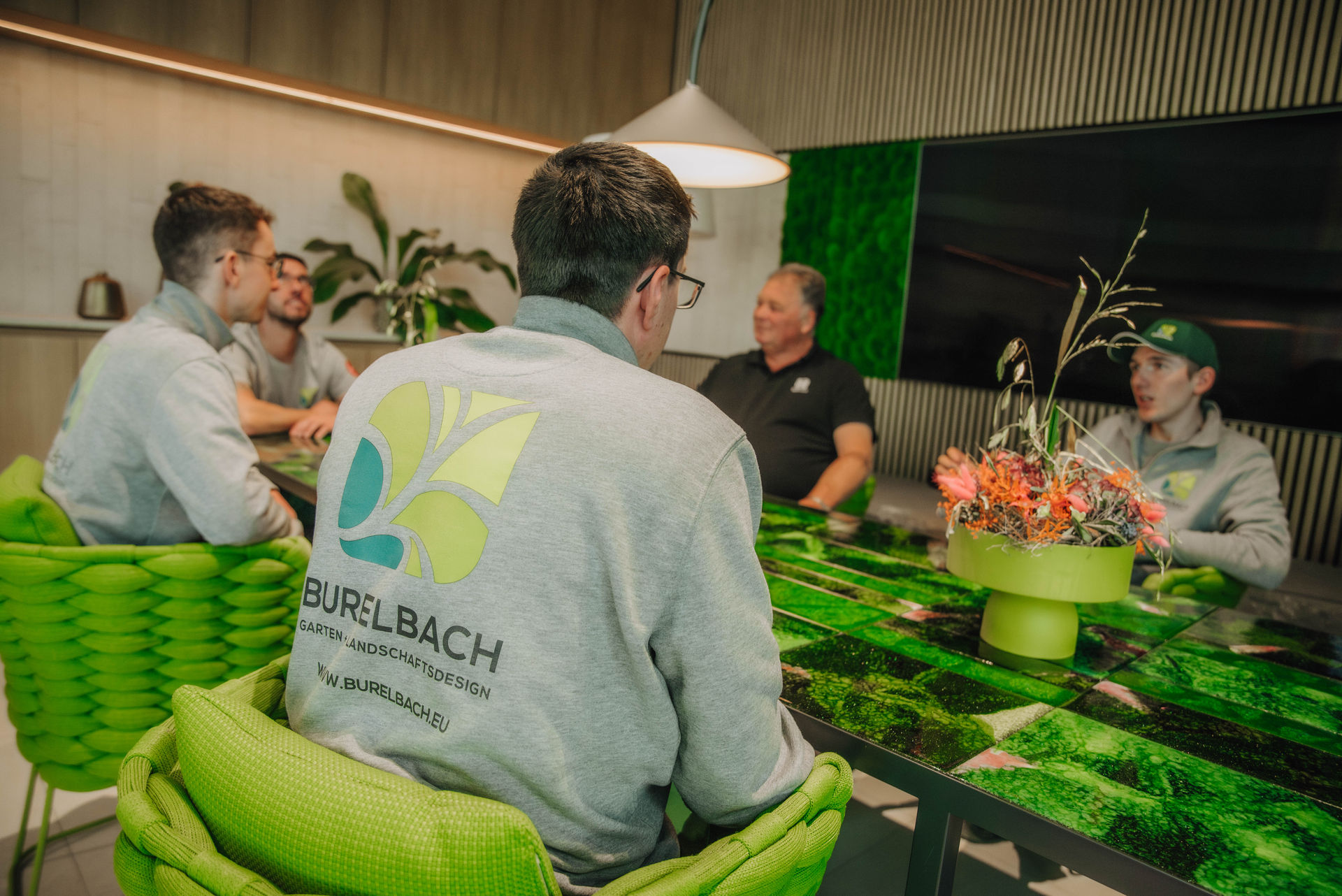 Fünf Personen sitzen um einen grünen, modernen Konferenztisch, einige tragen Sweatshirts der Marke BURELBACH. Der Raum spiegelt ihre Expertise im Garten- und Landschaftsbau wider, mit üppigen grünen Akzenten und einem stilvollen Pflanzenarrangement, das zum Gardendesign inspiriert.