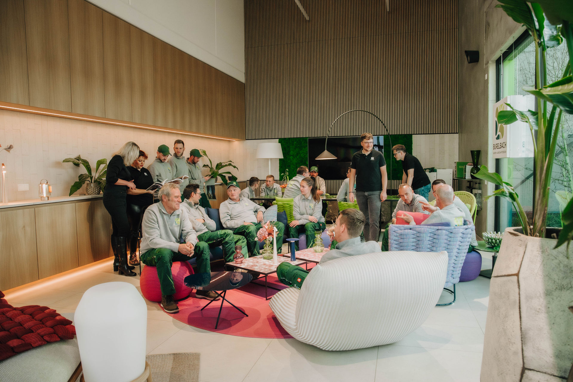 In einer modernen, hell erleuchteten Lounge mit stilvollen Gartenmöbeln, üppigen Pflanzen, einem Küchenbereich und bunten Möbeln von Burelbach sitzt und steht eine Gruppe von Menschen in legerer Kleidung und unterhält sich und unternimmt etwas.