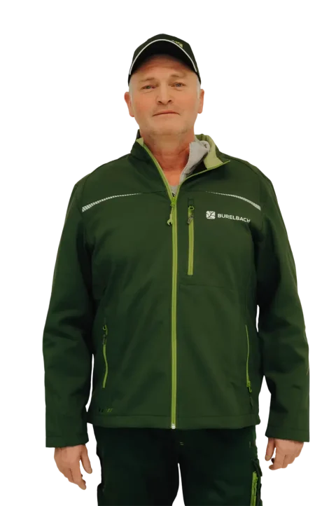Ein Mann mit einer dunkelgrünen Burelbach-Jacke und einer schwarzen Mütze steht mit dem Gesicht nach vorne. Die Jacke hat leuchtend grüne Reißverschlüsse, ein "Burelbach Garten- und Landschaftsbau"-Logo auf der Brust, und der Hintergrund ist transparent.
