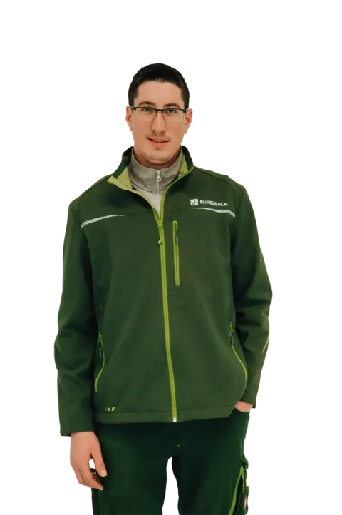 Ein Mann mit Brille und grüner Jacke mit dem Logo der Firma Garten- und Landschaftsbau steht mit dem Gesicht nach vorne und lächelt leicht. Der Hintergrund ist weiß und die Person ist zentriert im Bild.
