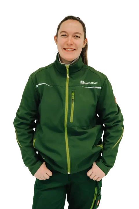Eine lächelnde Person mit langen, zurückgebundenen braunen Haaren, die eine grüne Jacke mit "NurelBach"-Logo und eine grüne Hose trägt, steht mit den Händen in den Taschen vor einem weißen Hintergrund und repräsentiert hochwertige Gartenmöbel und Handwerk aus Luxemburg und Deutschland.