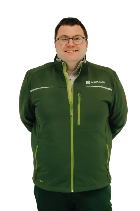 Eine lächelnde Person mit kurzen dunklen Haaren und Brille trägt eine grüne Jacke mit einem "Burkbach"-Logo und steht mit den Händen auf dem Rücken vor einem schlichten Hintergrund, der das Handwerk im Garten- und Landschaftsbau in Luxemburg und Deutschland repräsentiert.