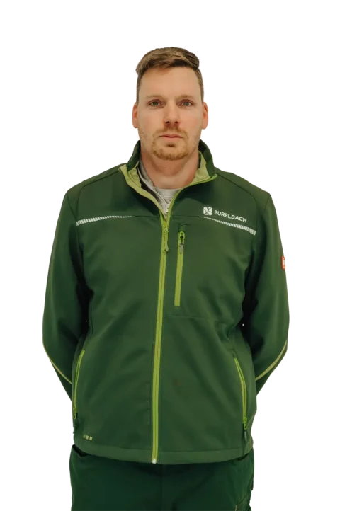 Ein Mann, der eine dunkelgrüne Jacke mit lindgrünen Reißverschlüssen und einem "Burelbach"-Logo trägt, bekannt für Gardendesign in Luxemburg und Deutschland, steht mit den Händen auf dem Rücken vor einem unscharfen Hintergrund.