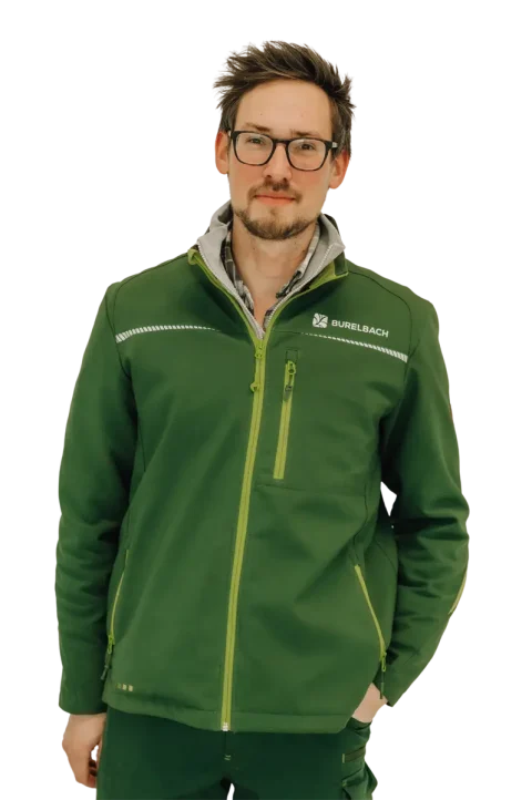 Ein Mann mit Brille und Gesichtsbehaarung trägt eine grüne BURELBACH Jacke mit Reißverschluss. Er steht mit neutraler Miene vor einem weißen Hintergrund und verkörpert den nachdenklichen Stil, der oft im modernen Gardendesign zu finden ist.