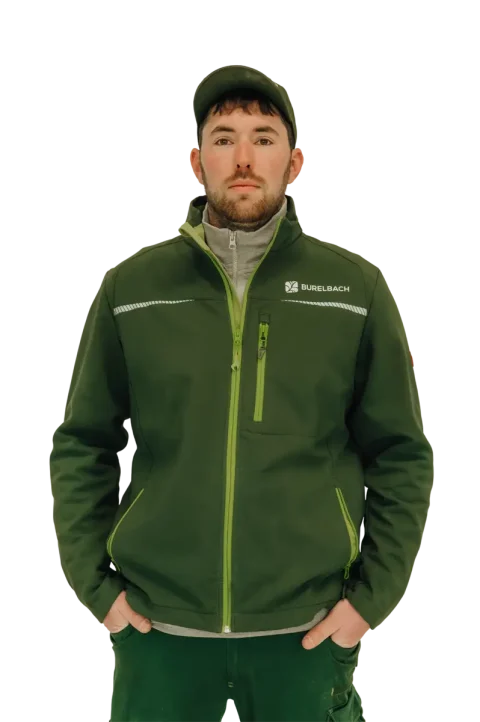 Ein Mann, der eine grüne Burelbach-Jacke, eine grüne Hose und eine grüne Mütze trägt, steht mit den Händen in den Jackentaschen nach vorne gerichtet. Der Hintergrund ist weiß. Burelbach ist in Luxemburg und Deutschland für seinen Stil bekannt.