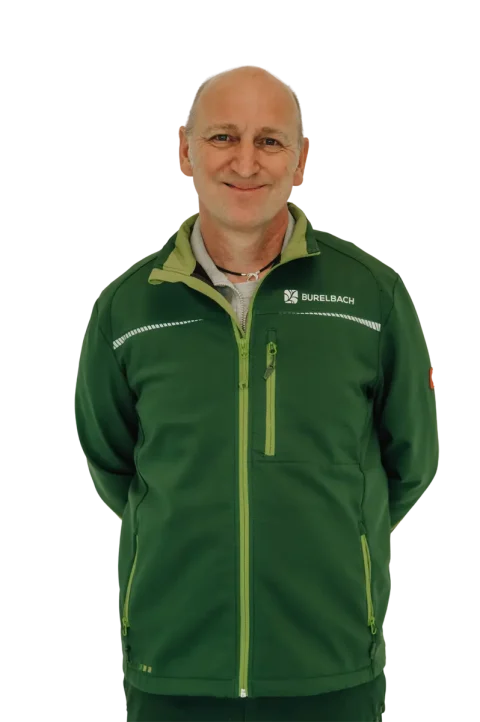 Ein lächelnder Mann in einer grünen Jacke mit dem "BURELBACH"-Logo, bekannt für seine Kompetenz im Garten- und Landschaftsbau, steht mit den Händen auf dem Rücken vor einem weißen Hintergrund.