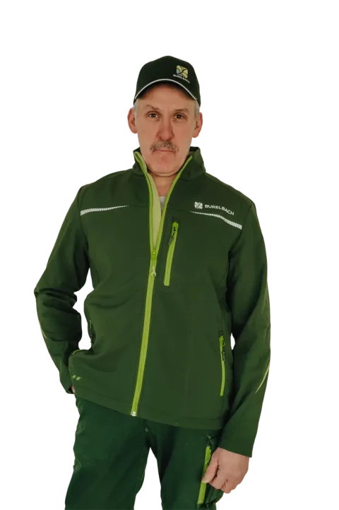 Ein Mann, der eine grüne Jacke mit leuchtend grünen Reißverschlüssen und eine dazu passende Mütze trägt, beide mit dem Logo von Burelbach Garten- und Landschaftsbau versehen, steht mit einer Hand in der Tasche vor einem weißen Hintergrund.