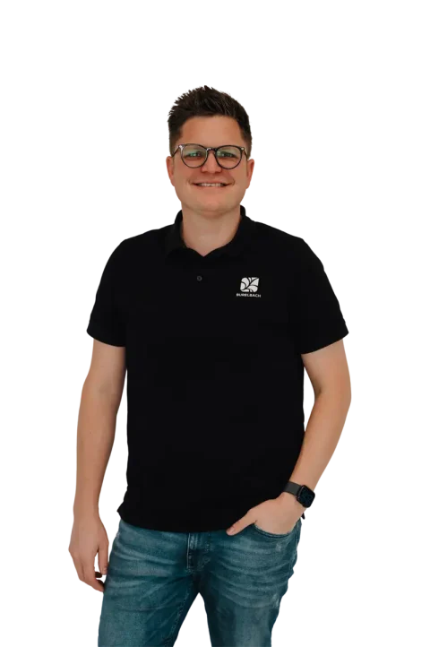 Eine lächelnde Person mit Brille, schwarzem Polohemd mit weißem Logo und blauer Jeans steht vor einem unscharfen Hintergrund und hat die linke Hand in der Tasche. Sie repräsentiert die Gardendesign-Trends in Luxemburg und Deutschland.