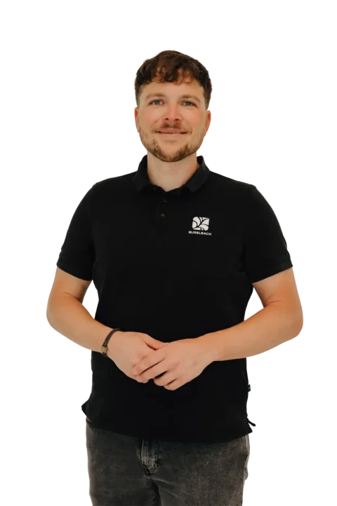 Ein Mann mit kurzen braunen Haaren und Bart lächelt, trägt ein schwarzes Polohemd mit weißem Logo auf der Brust und dunkle Jeans. Er steht vor einem schlichten weißen Hintergrund und ist bereit, sein handwerkliches Fachwissen über Gartenmöbel für Luxemburg und Deutschland weiterzugeben.