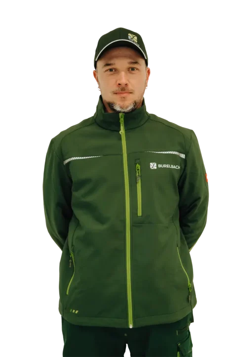 Ein Mann mit grüner Jacke und passender Mütze mit dem von Gardendesign und Handwerk inspirierten "BURELBACH"-Logo steht mit dem Gesicht nach vorne, die Hände hinter dem Rücken, vor einem weißen Hintergrund.