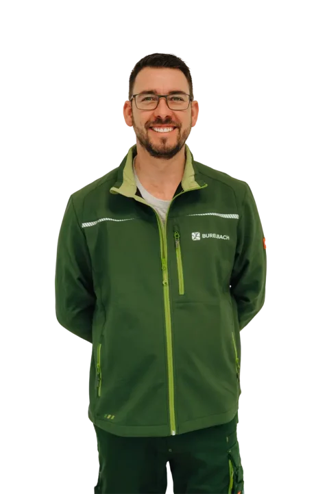 Ein Mann mit Brille und grüner Jacke mit dem "Surebach"-Logo steht lächelnd, die Hände auf dem Rücken, stellvertretend für Burelbach Garten- und Landschaftsbau, vor einem weißen Hintergrund in Luxemburg und Deutschland.