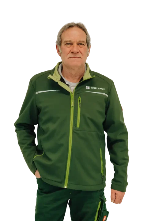 Ein Mann mittleren Alters mit kurzen grauen Haaren steht mit dem Gesicht nach vorne und trägt eine grüne Jacke mit einem "Burelbach"-Logo und eine grüne Hose, die das Handwerk in Luxemburg und Deutschland repräsentiert. Der Hintergrund ist unscharf mit Erdtönen.