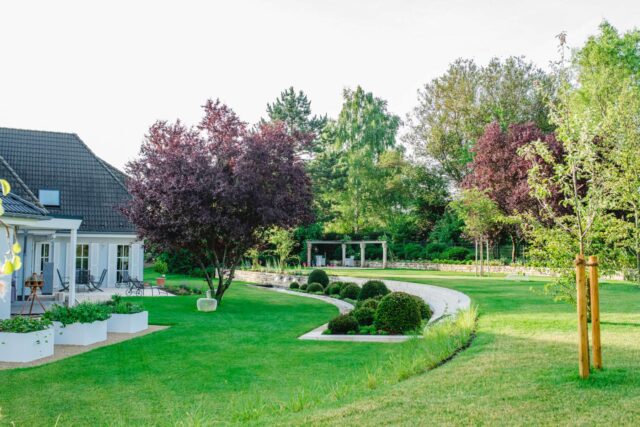Ein gepflegter Garten von Burelbach Gardendesign mit üppig grünem Rasen, ordentlich geschnittenen Sträuchern, einem geschwungenen Steinweg und alten Bäumen grenzt an ein modernes weißes Haus mit einer Terrasse und einem Sitzbereich im Freien - ein wahres Meisterwerk des Handwerks.