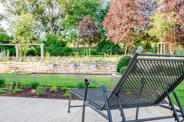 Ein schwarzer Metall-Loungesessel von Burelbach Handwerk blickt auf einen begrünten Garten mit einer Steinmauer, kleinen Springbrunnen, grünem Gras, blühenden Blumen und Bäumen im Hintergrund unter einem klaren Himmel.