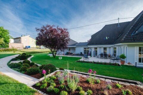 Ein gepflegter Garten mit grünem Rasen, Blumenbeeten, Sträuchern und Bäumen umgibt ein modernes weißes Haus mit Ziegeldach und Terrassenbereich, in dem stilvolle Gartenmöbel stehen - ein Beispiel für hervorragenden Garten- und Landschaftsbau in Luxemburg und Deutschland.