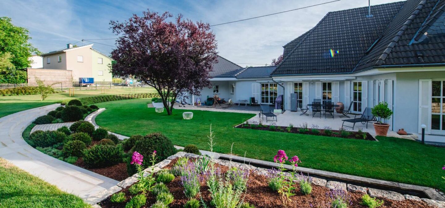 Modernes Haus mit einem großen Innenhof mit eleganten Gartenmöbeln, Sitzgelegenheiten im Freien und einem gepflegten Garten. Der geschwungene Steinweg inmitten von alten Bäumen und üppigen Rasenflächen in Luxemburg und Deutschland zeugt von hoher handwerklicher Qualität.
