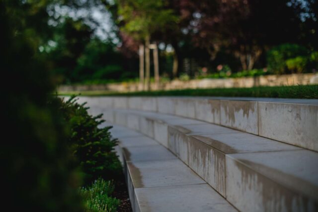 Geschwungene Steintreppen in einem Park, umrahmt von Grün und Bäumen, mit nassen Flecken, die auf kürzlichen Regen hindeuten. Die Szene zeigt fachkundigen Garten- und Landschaftsbau vor einem verschwommenen Hintergrund aus üppigem Laub.