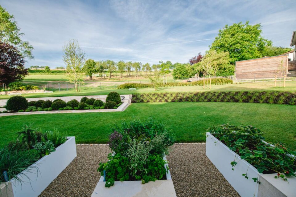Ein angelegter Garten mit sauber getrimmtem Gras, Pflanzkästen mit Kräutern, Sträuchern und kleinen Bäumen zeigt den handwerklichen Garten- und Landschaftsbau. Im Hintergrund sind offene Felder, Bäume und ein Holzgebäude unter blauem Himmel zu sehen.