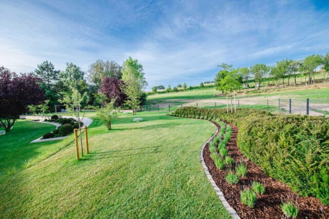 Ein gepflegter Garten von Burelbach zeigt fachmännisches Gardendesign mit gemähtem grünen Gras, geschwungenen Blumenbeeten, jungen Bäumen und einer Kulisse aus sanften Hügeln und verstreuten Bäumen unter einem klaren blauen Himmel.