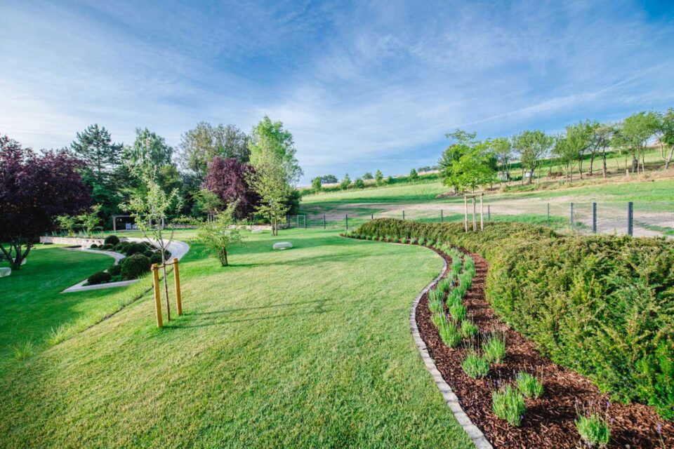 Ein gepflegter Garten von Burelbach zeigt fachmännisches Gardendesign mit gemähtem grünen Gras, geschwungenen Blumenbeeten, jungen Bäumen und einer Kulisse aus sanften Hügeln und verstreuten Bäumen unter einem klaren blauen Himmel.