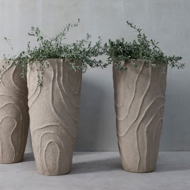 Drei hohe, strukturierte Steintöpfe mit wirbelnden Mustern beherbergen grüne Pflanzen vor einer glatten grauen Wand und einem grauen Boden - ein eleganter Hauch von Handwerk für Ihr Gartenmöbel-Ensemble.