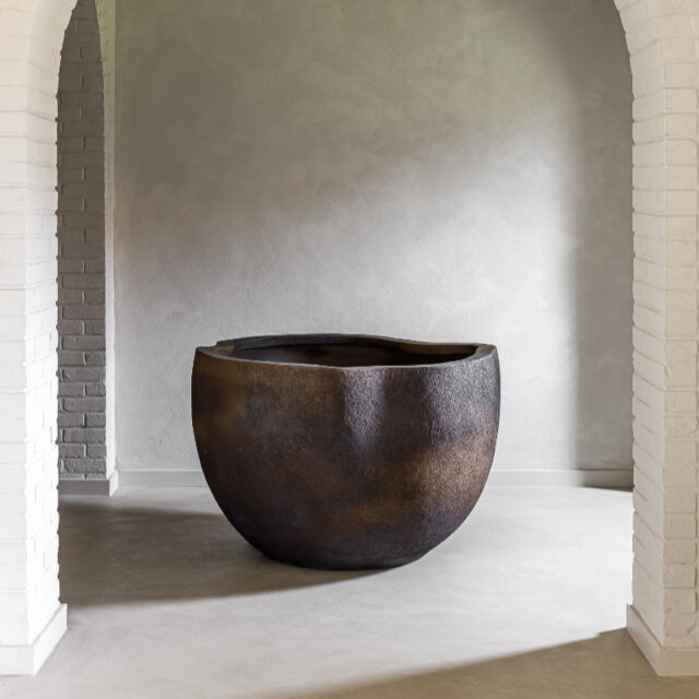 Ein großes, abgerundetes Waschbecken aus dunklem Stein steht auf einem glatten Boden, eingerahmt von zwei weißen Ziegelsteinbögen vor einer hellgrauen, strukturierten Wand in einem minimalistischen Interieur, das von Deutschland und Luxemburg inspiriertes Handwerk zeigt.