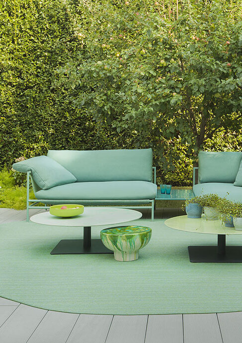 Eine moderne Sitzecke im Freien von Burelbach Garten- und Landschaftsbau besteht aus mintgrünen Sofas, passenden runden Tischen, einem gemusterten Hocker und einem runden Teppich auf einer Terrasse mit üppig grünen Bäumen und Sträuchern im Hintergrund.