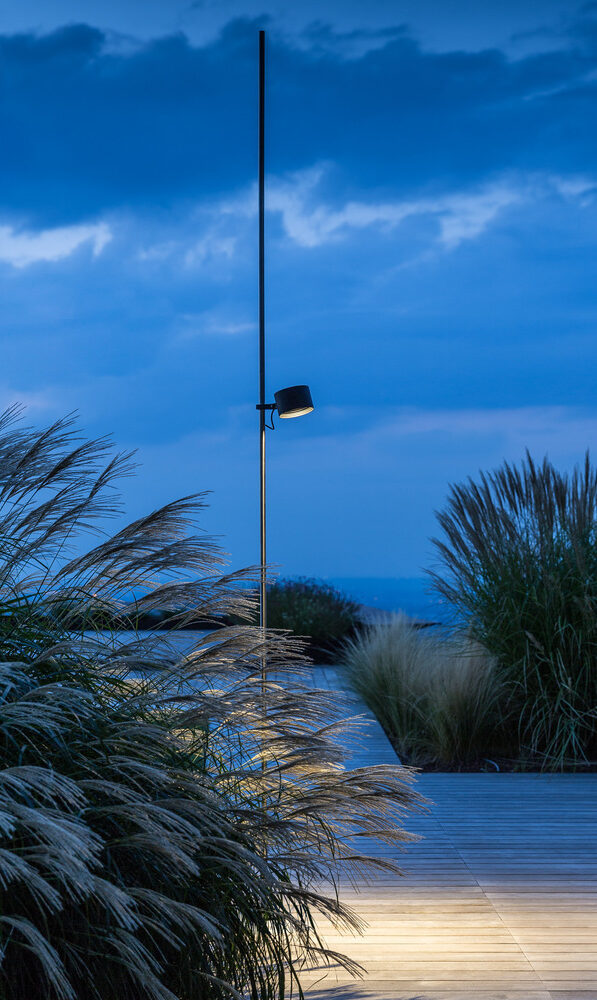 Eine moderne Außenleuchte, in der sich exquisites Handwerk widerspiegelt, beleuchtet einen von hohen Gräsern umgebenen Holzweg - eine elegante Note für Gardendesign in Deutschland und Luxemburg, vor einem blauen Abendhimmel mit Wolken.