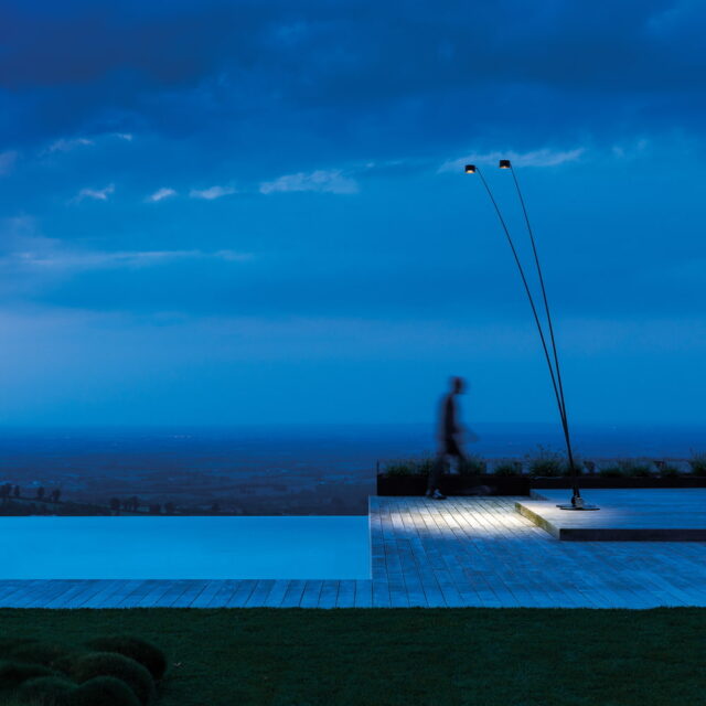 Eine Person geht in der Abenddämmerung neben einer modernen Poolterrasse spazieren. Zwei hohe, geschwungene Außenlampen von Burelbach beleuchten den Bereich, und unter dem stimmungsvollen blauen Himmel ist eine weitläufige Landschaft zu sehen, die vom Experten Gardendesign gestaltet wurde.