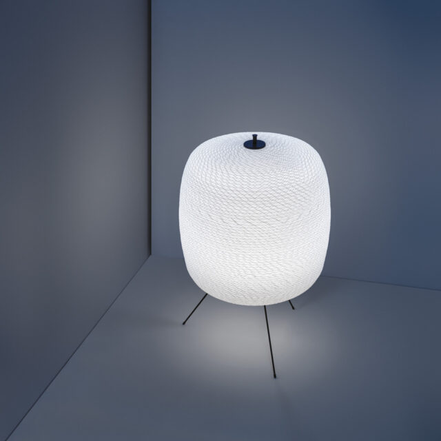 Eine moderne Lampe mit einem strukturierten, ovalen, weißen Schirm steht auf drei dünnen, schwarzen Beinen in einem minimalistischen Raum mit grauen Ecken. Perfekt, um Burelbach gartenmöbel zu akzentuieren, strahlt sie ein weiches Licht aus, das eine ruhige und moderne Atmosphäre schafft.