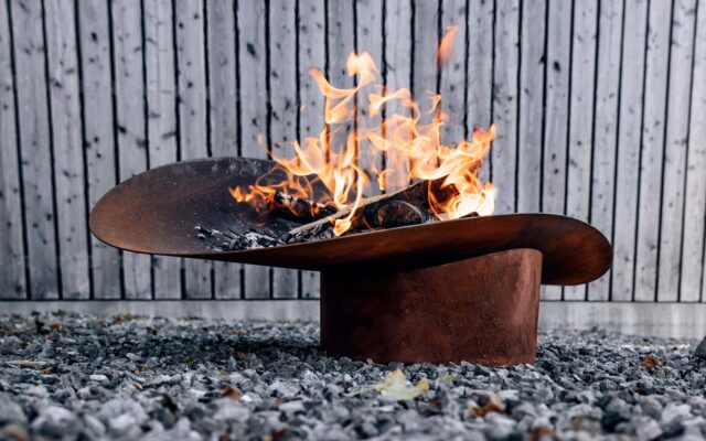 Eine moderne, skulpturale Feuerstelle aus Metall mit hellen Flammen, die auf Kies vor einem vertikalen Holzzaun steht - perfekt für alle, die Gardendesign und fachmännisches Handwerk schätzen.