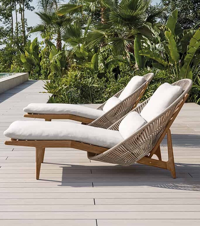 Zwei gepolsterte Loungesessel mit Holzrahmen und geflochtenen Details von Burelbach Handwerk stehen nebeneinander auf einem hellen Deck, umgeben von üppig grünen tropischen Pflanzen - ein perfekter Touch für inspiriertes Gartendesign.