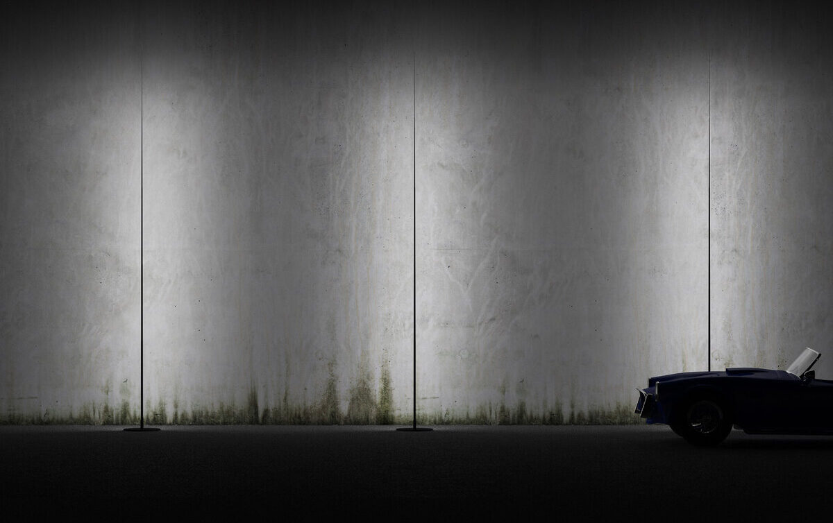 Eine dunkle, minimalistische Szene mit einer Betonwand, die von drei hohen, schmalen Leuchten beleuchtet wird, bildet die Kulisse. Ganz rechts parkt ein dunkelblaues Cabrio, das exquisites Gardendesign zeigt, meist im Schatten, nur die Front ist sichtbar.