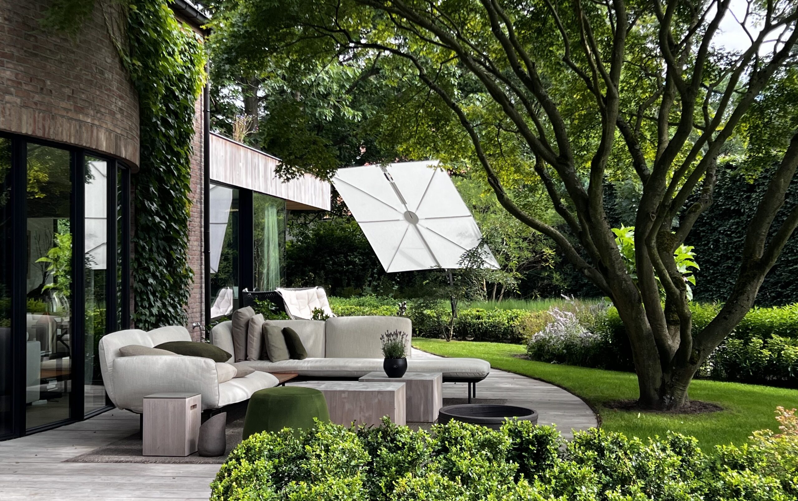 Eine moderne Außenterrasse mit stilvollen Gartenmöbeln, einem geschwungenen Sofa, Kissen und kleinen Tischen liegt auf einer Holzterrasse inmitten von üppigem Grün, perfekt für ein anspruchsvolles Gardendesign neben einem Haus mit Glaswänden.