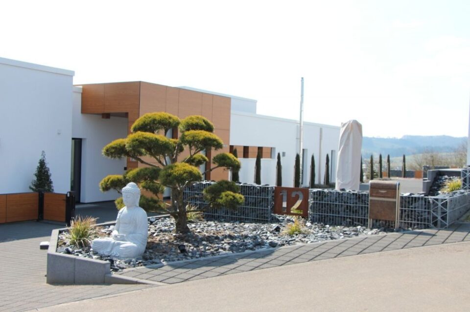 Ein modernes weißes Gebäude mit hölzernen Akzenten von Burelbach, begrünt mit Felsen, Garten- und Landschaftsbauelementen, einem beschnittenen Baum und einer weißen Buddha-Statue davor; die Zahl 12 steht auf einem Metallschild neben dem Eingang.